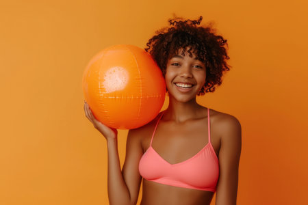 Funny young happy African American woman in swimsuit having fun holding inflatable ball and going on summer holiday trip standing on orange studio background. Vacation tour and travel conceptの素材