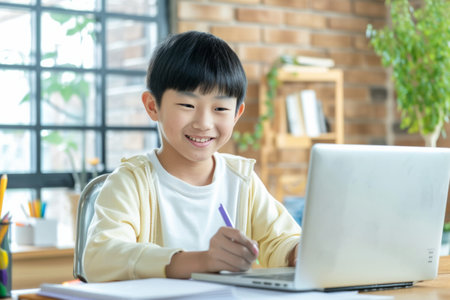 Smiling Japanese Boy studying with laptop computer. Teenage boy sitting at his desk and writing in notebook. Student doing his homework or learning online.の素材