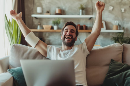 Man with an arm injury relaxing at home. Happy man with a sling on his broken arm sitting on the sofa, watching a funny movie on a laptop computer and laughing.の素材