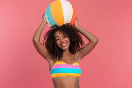 Funny young happy African American woman in swimsuit having fun holding inflatable ball and going on summer holiday trip standing on pink studio background. Vacation tour and travel concept.の素材