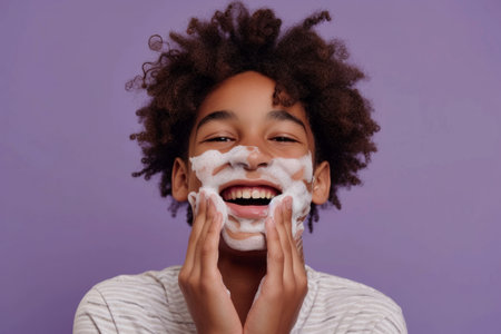 Handsome nervous African American teenage boy with foam on face learning to shave isolated on pink studio backgroundの素材