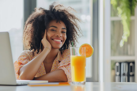 Funny happy smiling African American woman sitting at the desk on workplace at office with juice coctail and booking tickets for summer vacation online via laptop.の素材
