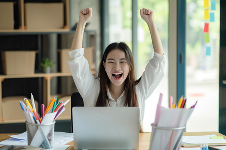 Happy excited young Japanese business woman accountant standing at the desk working on laptop computer with a pile of documents on table in office and making yes gesture rejoicing in successful job.の素材