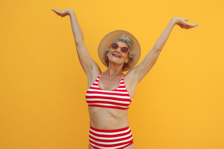 Cool funky elderly Latin American woman dressed striped red swimsuit dancing having fun isolated yellow color backgroundの素材