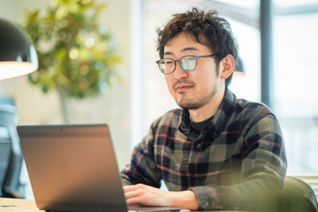 A motivated Japanese man concentrates on his laptop in a well-lit office, showing success and positivity in his work.の素材
