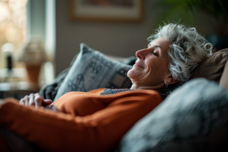 In the comfort of her living room, a serene and content mature woman lies on the sofa, embracing the tranquility of her lazy weekend at home. With a gentle smile, she dreams and breathes in the fresh air, feeling carefree and relaxed after a hectic working day. Resting on the comfortable couch, she enjoys the blissful pastime with joy.の素材
