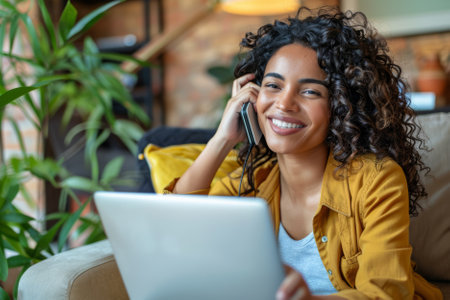 A cheerful Latina woman multitasks effortlessly, engaging in a phone conversation while diligently working on her laptop from the comfort of her home.の素材
