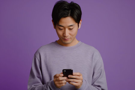 Against a calming purple background, an attentive Japanese man is typing a text message on a smartphone app, maintaining his online presence with ease and efficiency.の素材