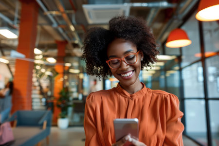 A joyful African American businesswoman utilizing her smartphone in a vibrant, contemporary office setting.の素材
