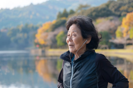 A Japanese senior woman listening to music while running by the lake in nature. The elderly woman is exercising to stay healthy, vital, enjoying physical activity and relaxation outdoors.の素材