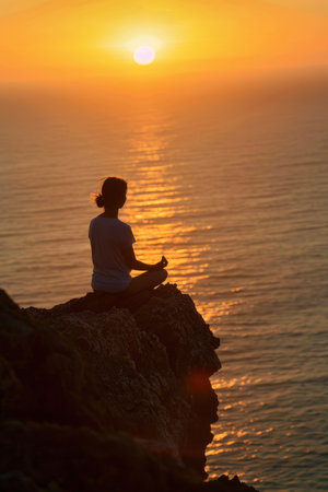 Silhouette of a woman meditating on a cliff at sunsetの素材