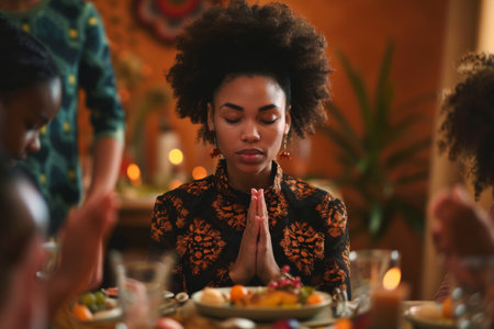 a Black Woman Saying Grace at Dinner: A Black woman standing at the dinner table with her family, saying grace before a meal, her eyes closed and hands folded in prayer. The studio background is a warm orange, creating a cozy atmosphereの素材