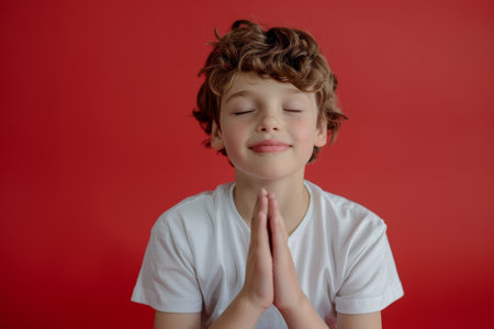 a young Caucasian boy praying with his hands folded: He sits with his hands folded in his lap, eyes closed and a peaceful expression on his face on a red studio backdropの素材
