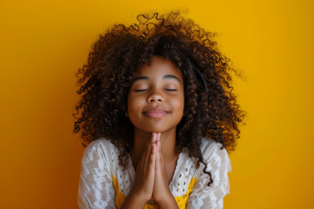 a young African American girl praying with her hands clasped: She kneels with her hands clasped and eyes closed, a serene smile on her face on a bright yellow studio backdropの素材