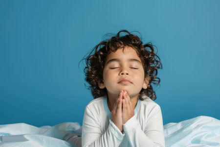 a Hispanic Child Saying Bedtime Prayers: A young Hispanic child kneeling by their bed, saying bedtime prayers with closed eyes and folded hands. The studio background is a soothing blue, evoking a sense of peace and securityの素材