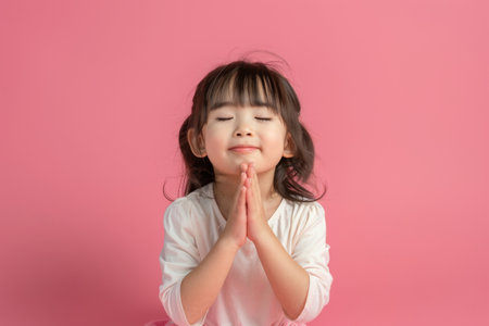 a young Japanese girl praying with her hands clasped: She kneels with her hands clasped in front of her, eyes closed and a serene smile on her face on a pink studio backgroundの素材