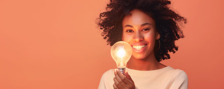 A young woman holding a softly glowing light bulb, isolated on a peach studio background. The light highlights her gentle smile.の素材