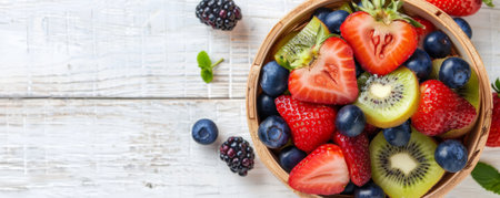 A top-down view of a wooden basket filled with nutritious fruits such as strawberries, blueberries, and kiwis, with ample text space on a light wooden surface.の素材
