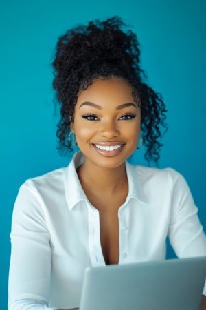 Young African American female entrepreneur working on her business project, smiling brightly at the camera while typing on a laptop, in a minimalist office with a blue background.の素材