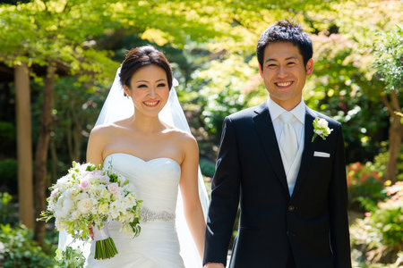 A happy Japanese bride and groom walking through a garden after their wedding ceremony, holding hands and smiling at the camera.の素材