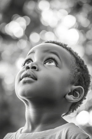 Black and white portrait of a preschooler African American boy with short hair, expressive eyes, and clear skin, looking up in natural daylight.の素材