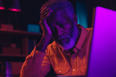 Senior African American man with a beard, anxiously clutching his head at a desk, dark purple lighting.の素材