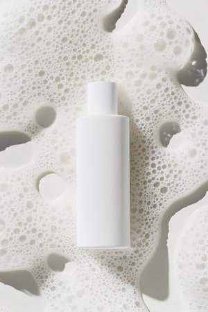 A close-up top view of a simple facial cream bottle, surrounded by smooth, shampoo-like bubbles. The minimal design of the bottle is highlighted by the shiny, airy texture of the surrounding foam.の素材