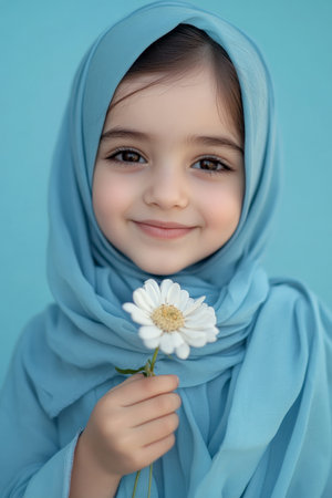School-age Arabian girl smiling shyly with a grateful expression, her small hands gently holding a flower, against a soft blue background.の素材