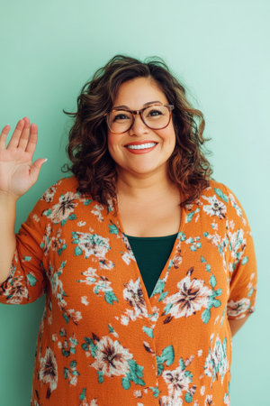 Middle-aged Hispanic woman with glasses, friendly smile, cheerful demeanor, standing against a clean light green background, showing warm and approachable feelings, slightly waving.の素材