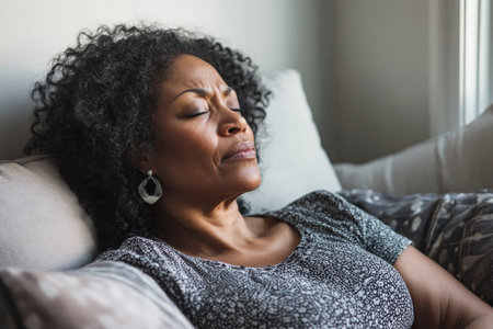 A middle-aged African American woman reclines on a couch, her expression strained as she struggles with a headache. The clean, neutral background emphasizes her discomfort, highlighting the challenges of balancing life while feeling unwell.の素材