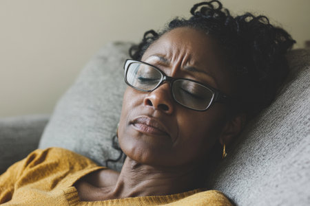 A middle-aged African American woman reclines on a couch, her expression strained as she struggles with a headache. The clean, neutral background emphasizes her discomfort, highlighting the challenges of balancing life while feeling unwell.の素材