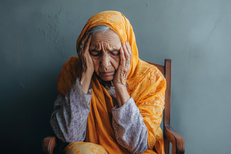 An elderly Arabian woman sits in a chair, holding her head and looking exhausted. The minimalist background emphasizes her discomfort, capturing the poignant reality of experiencing physical discomfort as one ages.の素材