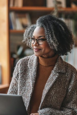 A sophisticated middle-aged African American woman stands in her home office, listening to inspiring music while working on her laptop. Her confident smile and focused demeanor show her passion for both her career and the uplifting sounds that fuel her creativity.の素材