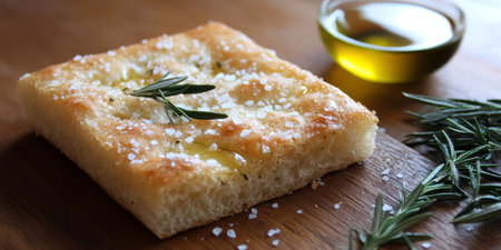 A traditional loaf of Italian focaccia bread, topped with rosemary and sea salt, rests on a wooden table, alongside olive oil for dippingの素材