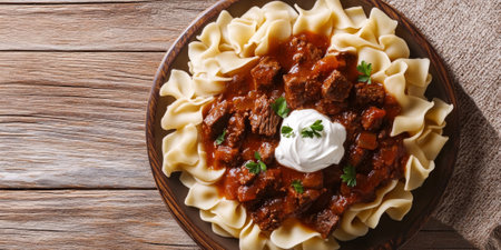 A hearty Hungarian goulash, with tender beef, onions, and paprika-spiced sauce, served with buttered egg noodles and a dollop of sour cream on a rustic wooden table. The dish feels warm and filling.の素材