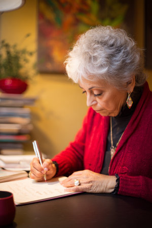 An elderly Hispanic woman writes personalized holiday cards to corporate partners. Her thoughtful traditions nurture long-lasting business connections.の素材