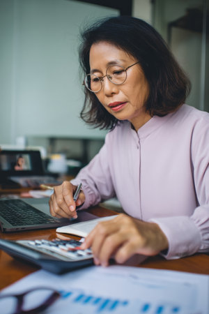 A middle-aged Asian woman modifies spreadsheet formulas while reviewing forecasts on a video call. Her accuracy supports financial stability.の素材
