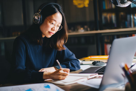 Young adult Asian woman attending a webinar about digital transformation, taking notes diligently with headphones on in a quiet workspaceの素材