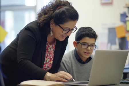 Mature Hispanic businesswoman leaning over a desk with a younger colleague, reviewing social media metrics together on a laptop, teamwork and mentorship in focusの素材