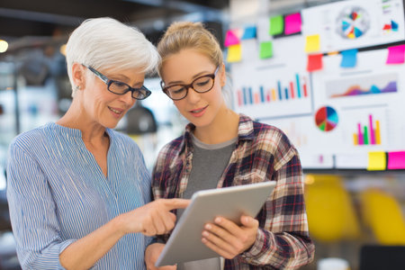 Mature Caucasian woman mentoring a junior analyst on data visualization techniques, pointing to colorful charts on a tabletの素材