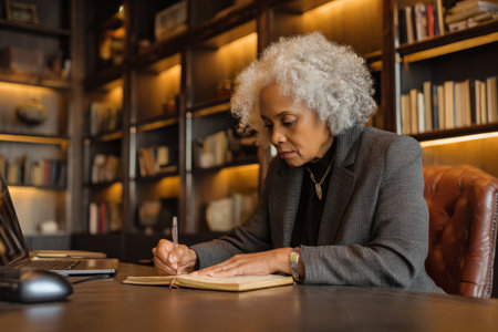 Elderly African-American business woman attending an executive board meeting via video conference, taking notes in a leather-bound journal, wearing an elegant dark gray blazer, office lined with bookshelves and soft lighting, creating a professional yet personal spaceの素材