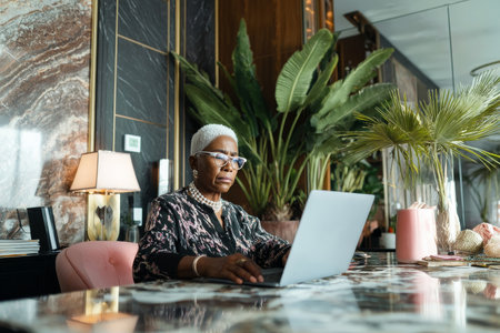 An elderly African-American woman works on financial forecasts at her marble desk, glasses on, surrounded by motivational art and a leafy plant.の素材