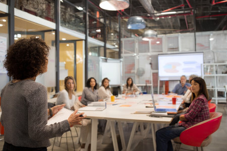 A mature Hispanic woman in a modern open-plan office leads a diversity training workshop, using slides and real-life case studies to highlight inclusive leadership, empathy, and cultural awareness among employees. Everyone is engaged and taking notes attentivelyの素材