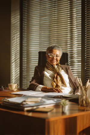 Elderly African-American business woman sitting confidently at a modern wooden office desk, surrounded by documents and a cup of tea, wearing a stylish scarf and glasses, with sunlight pouring through the window blinds in the backgroundの素材