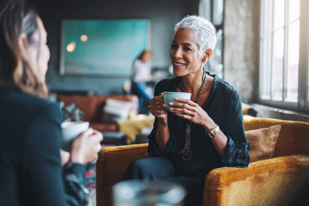 A mature Black businesswoman chats over coffee with a colleague in a relaxed office lounge, exchanging thoughts on mentorship and career progress.の素材