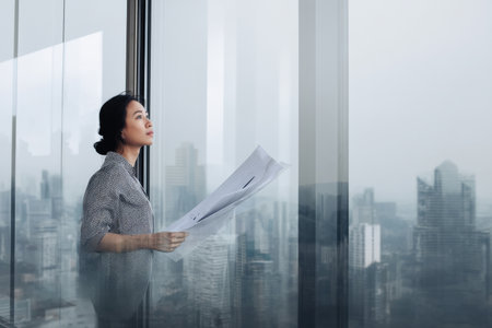A middle-aged Asian woman reviews printed analytics in a sleek glass office, framed by a city skyline that hints at growth, ambition, and her leadership vision.の素材