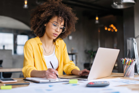 A young adult African-American woman in a bright coworking space updates financial reports on her laptop while on a video call with her manager, discussing budget forecasts and quarterly revenue goals with confidence and professionalismの素材