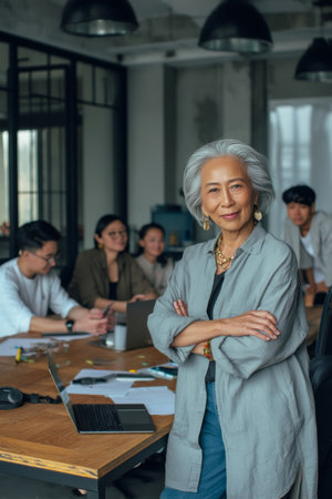 Asian elderly businesswoman leaning on a conference table during a brainstorming session, hands crossed with a confident smile, surrounded by younger colleagues with laptops, collaborative mood and modern industrial decorの素材