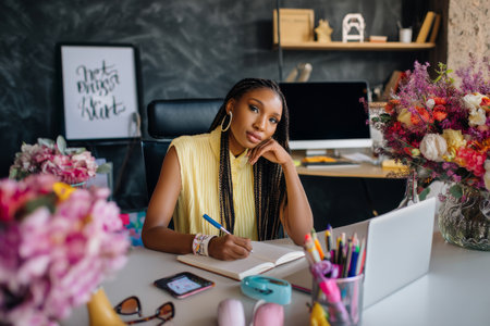 A young Black woman with braids writes in a notebook at her lively desk, surrounded by tech, flowers, and inspiring quotes in a bright creative space.の素材