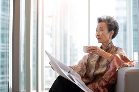 Asian elderly business woman relaxing in a modern office lounge area, sipping coffee while reading a report, dressed in a luxurious silk blouse, soft light coming from large windows, reflecting her wisdom and calm demeanorの素材